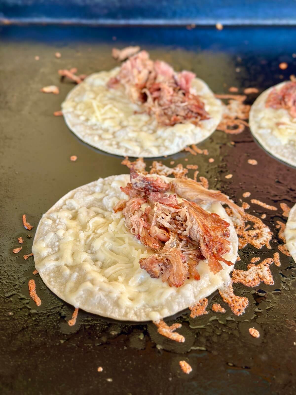 corn tortillas frying on a griddle with shredded cheese and pulled pork on top