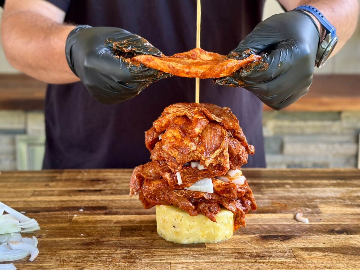 a thick slice of pineapple with a wooden skewer through it standing straight up, and a man placing thin slices of pork butt on the skewer