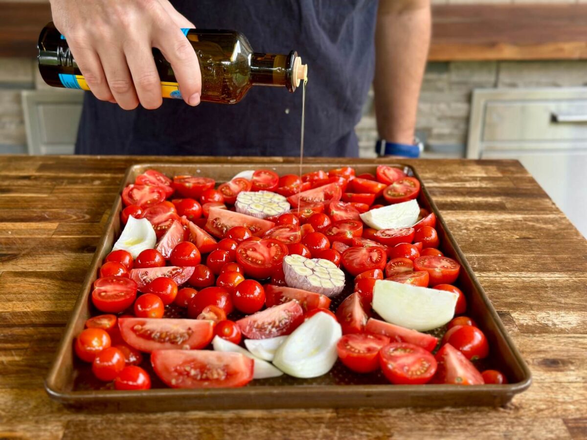 drizzling olive oil over a sheet tray of cut tomatoes, garlic, and onions