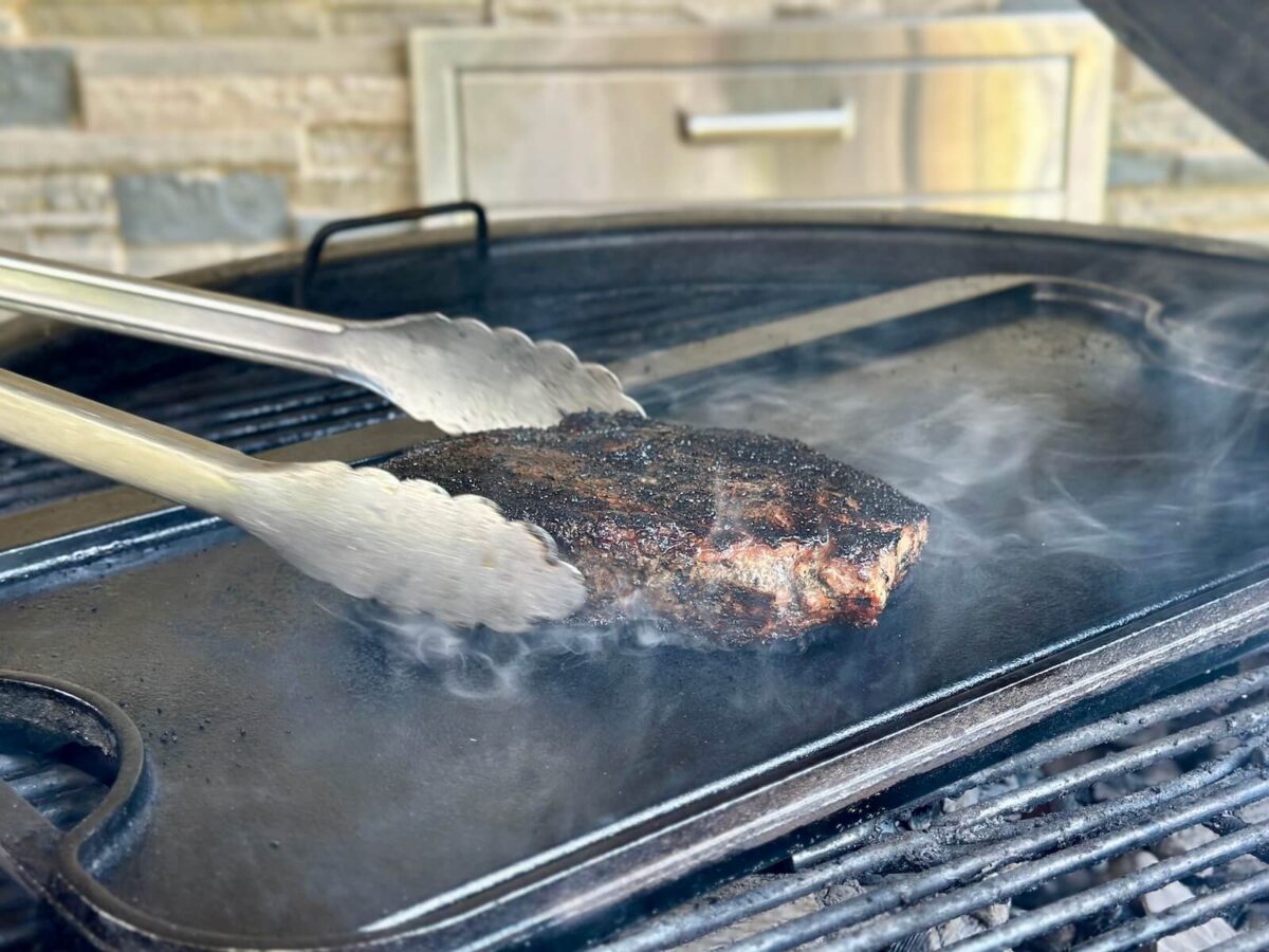 cooking a ribeye steak with a charred black crust on a cast iron griddle set on top of a kettle charcoal grill